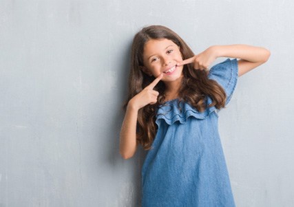 Young girl wearing blue dress and pointing to healthy smile   