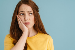 Woman on blue background holding her jaw and looking miserable